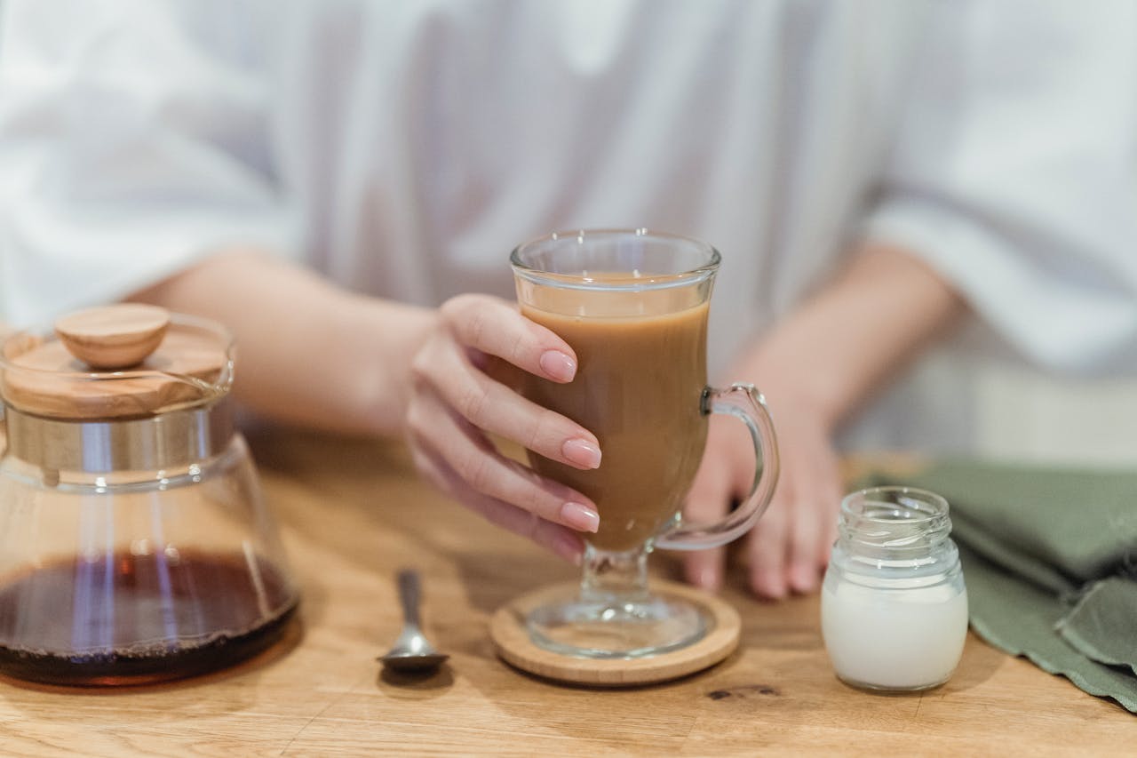 A close-up of a hand holding a coffee mug on a wooden table, with a pot and milk jar.
