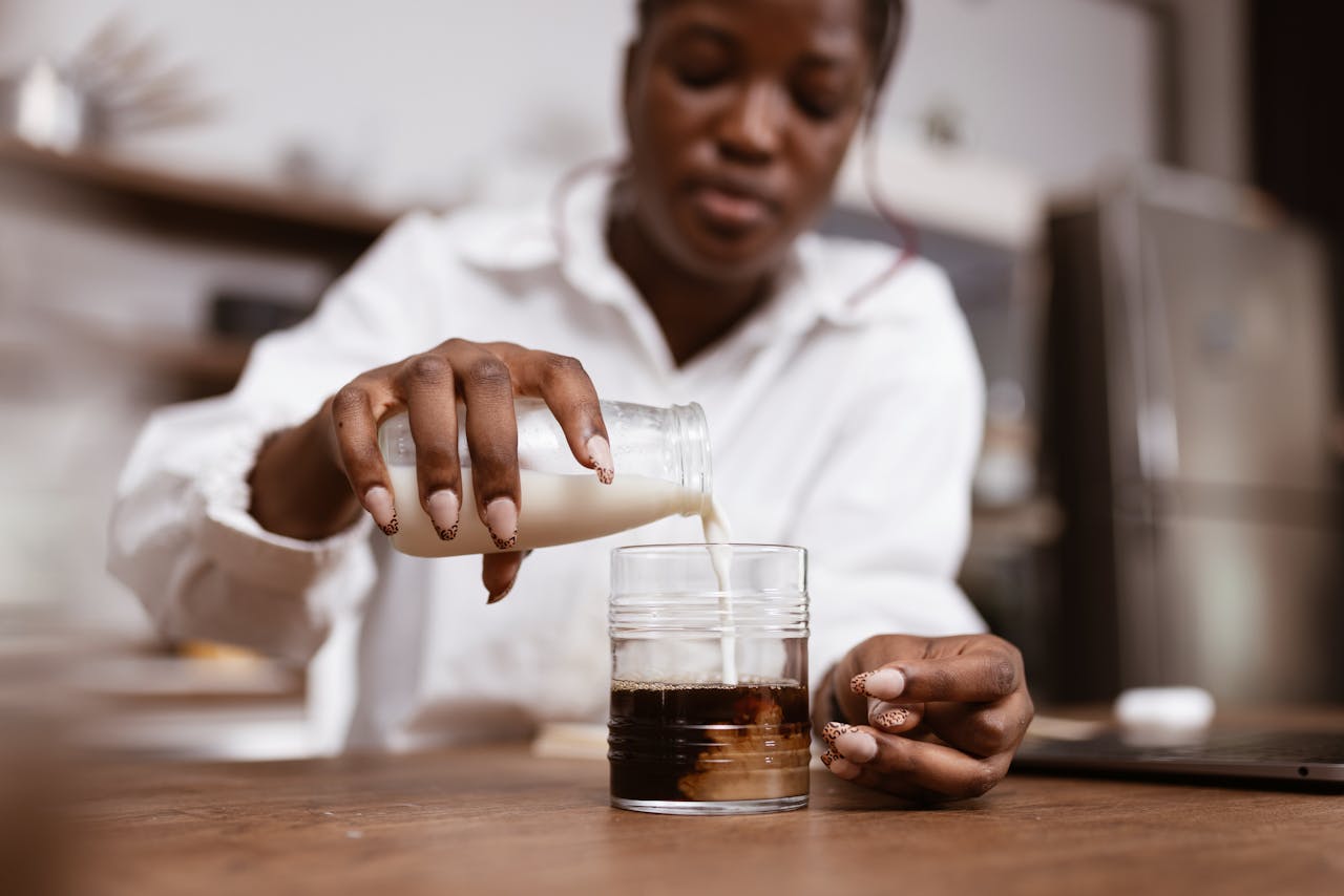 why-choose-us A woman pours milk into a glass of coffee on a wooden table, focused on brewing.